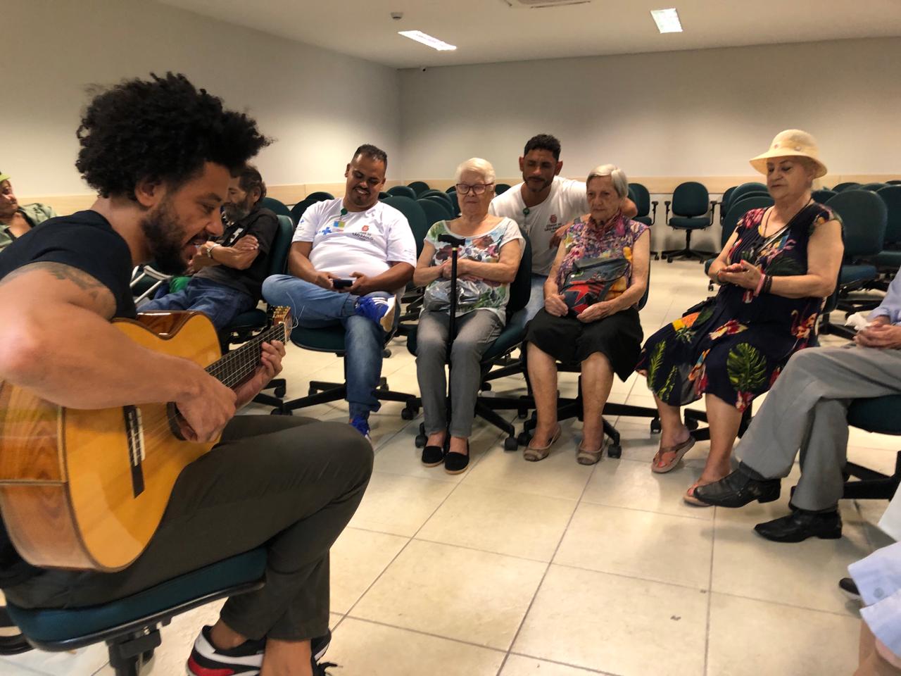 Na imagem, Leandro, um homem negro e alto, está sentado em uma cadeira tocando violão para um grupo de idosos